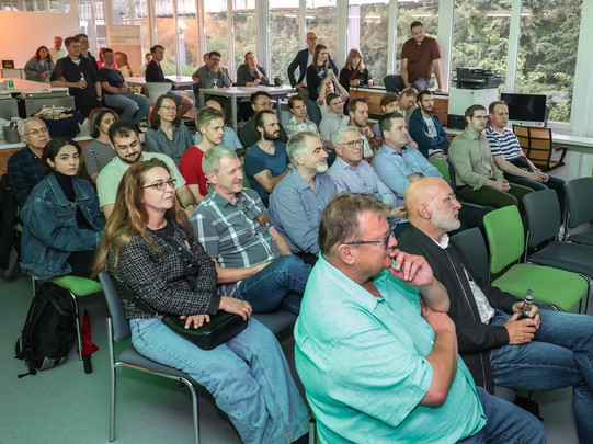 Foto: Im Vortragsraum sitzen Gäste in den vorderen Reihen, im Hintergrund stehen und sitzen weitere Personen.