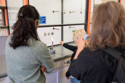 Zwei junge Frauen schauen einem blonden Kind zu, dass in einem Chemielabor etwas über einen Bunsenbrenner hält.