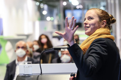 Eine Frau steht auf einem Podium und spricht ins Mikrofon. Dabei dreht sie sich leicht Richtung Kamera und hält die linke Hand hoch.