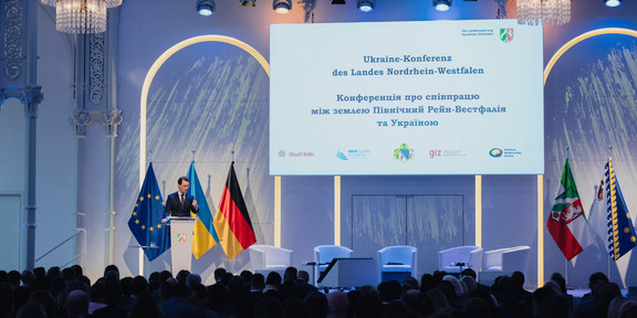 Ein Redner steht auf einer Bühne bei der Ukraine-Konferenz des Landes Nordrhein-Westfalen, mit EU-, Ukraine- und Deutschland-Flaggen im Hintergrund.