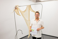 Im Zentrum des Bildes befindet sich Daniel Friedt, der mit seinem Preis-Zertifikat vor einer Kunstinstallation aus Plastik steht steht und in die Kamera lächelt. Im Hintergrund befinden sich weiße Wände.