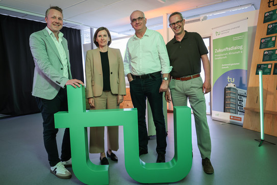 In the photo, you can see four people, including Prof. Manfred Bayer, standing behind the TU logo and smiling at the camera.