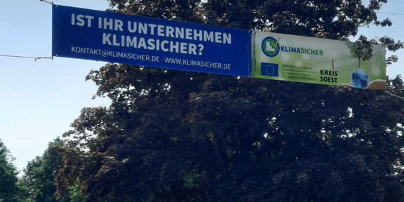 Banner des Projekts „KlimaSicher“ Ein Banner des Projekts „KlimaSicher“ ist über eine Straße in der Luft gespannt. Bäume und Häuser sind im Hintergrund.