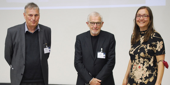 Auf dem Foto stehen drei Personen bei einer Veranstaltung nebeneinander und lächeln in die Kamera. Die beiden Männer (links) sind in Anzügen gekleidet, während die Frau (rechts) ein schwarz-rotes Kleid trägt.