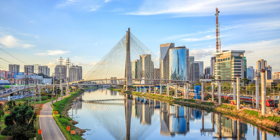 Foto: Die "Octavio Frias de Oliveira"-Brücke in São Paulo überspannt einen schimmernden Fluss. Neben der Brücke sind grüne Parks und glänzende Hochäuser mit Glasfronten zu sehen.