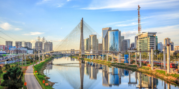 Foto: Die "Octavio Frias de Oliveira"-Brücke in São Paulo überspannt einen schimmernden Fluss. Neben der Brücke sind grüne Parks und glänzende Hochäuser mit Glasfronten zu sehen.