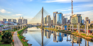 Foto: Die "Octavio Frias de Oliveira"-Brücke in São Paulo überspannt einen schimmernden Fluss. Neben der Brücke sind grüne Parks und glänzende Hochäuser mit Glasfronten zu sehen.