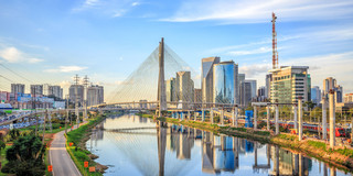 Foto: Die "Octavio Frias de Oliveira"-Brücke in São Paulo überspannt einen schimmernden Fluss. Neben der Brücke sind grüne Parks und glänzende Hochäuser mit Glasfronten zu sehen.