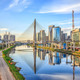Foto: Die "Octavio Frias de Oliveira"-Brücke in São Paulo überspannt einen schimmernden Fluss. Neben der Brücke sind grüne Parks und glänzende Hochäuser mit Glasfronten zu sehen.
