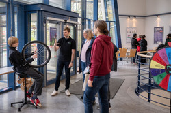 Ein Junge sitzt auf einem Stuhl und hält ein sich drehendes Rad zwischen den Händen. Um ihn stehen Menschen und ein Mann mit TU-T-Shirt, der etwas erklärt. Im Hintergrund sind weitere Besucher*innen an anderen Stationen zu sehen.