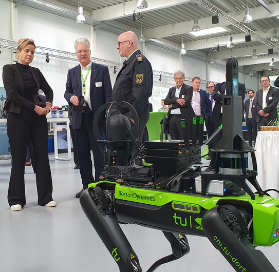 Exponat mit Menschen im Hintergrund Mona Neubaur, Prof. Wietfeld und Dirk Aschenbrenner von der Feuerwehr Dortmund und dem Deutschen Rettungsrobotikzentrum unterhalten sich und schauen auf ein roboterhundähnliches Exponat.