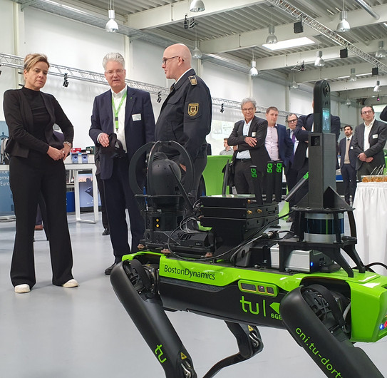 Exponat mit Menschen im Hintergrund Mona Neubaur, Prof. Wietfeld und Dirk Aschenbrenner von der Feuerwehr Dortmund und dem Deutschen Rettungsrobotikzentrum unterhalten sich und schauen auf ein roboterhundähnliches Exponat.