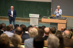 Zwei Männer sprechen vor einem vollbesetzten Hörsaal