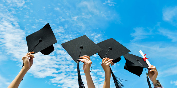 Four hands hold up black graduation caps. Blue sky can be seen in the background.