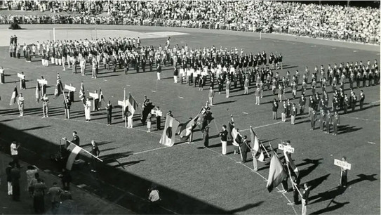 Black-and-white photograph of the opening ceremony of the “3rd International Week of University Sports” from 1953. Various athletes form formations on the pitch of the Rote Erde stadium. 