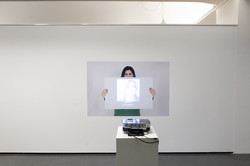 Ein Beamer wirft das Foto einer jungen Frau auf das hängende Bild einer anderen jungen Frau, die ein weißes Papier in die Höhe hält. 