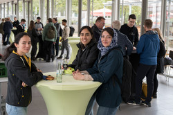 Drei junge Frauen in dunklen Jacken stehen um einen hellgrünen Stehtisch und lächeln in die Kamera. Im Hintergrund ein Empfang in einem Raum, in dem Menschen bei einem Empfang um Stehtische herum stehen.