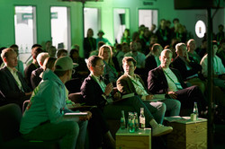 Foto: Mehrere Personen sitzen in einem grünen, modern beleuchteten Raum, während eine Person mit einem Mikrofon spricht.