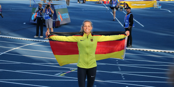 A smiling young woman in sportswear stands in a track and field stadium holding a German flag behind her back.