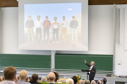 People in a lecture hall listen to the presentation of a man in a suit.