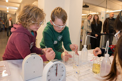 Das Bild zeigt einen Versuchstisch mit Stehuhren und verschiedenen Glasbehältern, die mit unterschiedlichen Substanzen gefüllt sind. Am Versuchstisch stehen zwei Kinder mit Schutzbrillen.