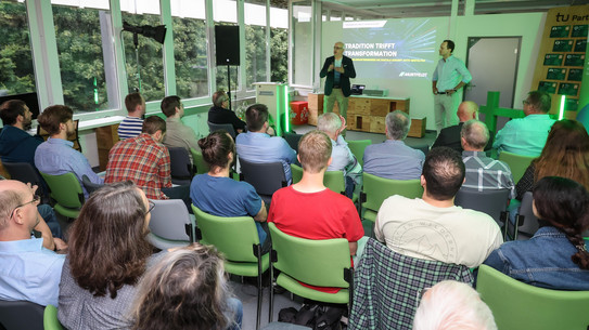 Foto: Redner steht vorne im Vortragsraum und spricht, im Vordergrund sind sitzende Gäste im Publikum.