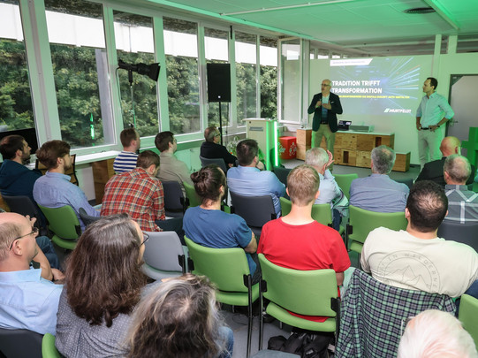 Foto: Redner steht vorne im Vortragsraum und spricht, im Vordergrund sind sitzende Gäste im Publikum.
