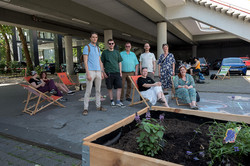 Photo: A group of people stands and sits near deck chairs and a planted raised bed under an overpass.