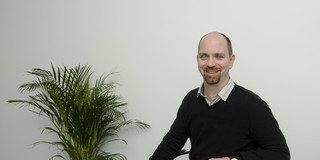 A man sits at a table next to a plant and smiles at the camera