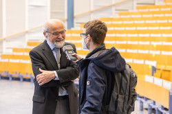 Vortragsreihe „Initialzündung“ Ein Mann im Anzug wird von einem Studenten in einem leeren Hörsaal interviewt.