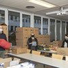 Drei Personen stehen in einem großen Raum voller Kartons mit gespendeten Lebensmitteln.