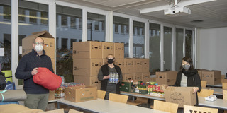 Drei Personen stehen in einem großen Raum voller Kartons mit gespendeten Lebensmitteln.