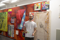 Ein Mann steht vor einer Wand an der Bilder in roten und gelben Farben einer Kunstausstellung hängen und hält eine Urkunde in den Händen.