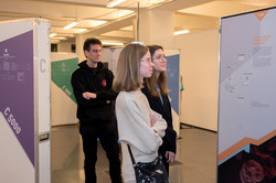 Das Bild zeigt zwei weibliche junge Erwachsene sowie einen männlichen Erwachsenen, die von Ausstellungswänden umgeben sind und sich diese anschauen.