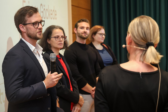 Foto: Ein Preisträger spricht ins Mikrofon, umgeben von seinen Mitpreisträger*innen.