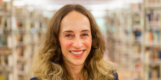 Porträtfoto: Prof. Katrin Röder steht slächelt und mit verschänkten Armen zwischen den Bücherregalen in einer Bibliothek. Sie trägt eine helle Bluse udn einen dunkelblauen Blazer.