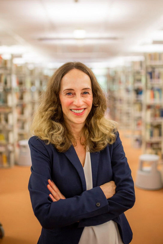Porträtfoto: Prof. Katrin Röder steht slächelt und mit verschänkten Armen zwischen den Bücherregalen in einer Bibliothek. Sie trägt eine helle Bluse udn einen dunkelblauen Blazer.