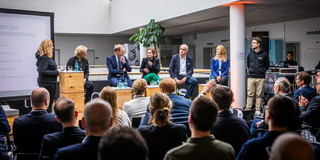 Im Vordergrund des Bildes ist ein Teil eines Publikums von hinten zu sehen. Vorne auf einer Erhöhung befinden sich FH-Rektorin Prof. Tamara Appel, IHK-Hauptgeschäftsführer Stefan Schreiber, die Geschäftsführerin der Wirtschaftsförderung Heike Marzen, TU-Rektor Prof. Manfred Bayer und ISM-Vizepräsidentin Prof. Audrey Mehn.