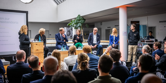 Im Vordergrund des Bildes ist ein Teil eines Publikums von hinten zu sehen. Vorne auf einer Erhöhung befinden sich FH-Rektorin Prof. Tamara Appel, IHK-Hauptgeschäftsführer Stefan Schreiber, die Geschäftsführerin der Wirtschaftsförderung Heike Marzen, TU-Rektor Prof. Manfred Bayer und ISM-Vizepräsidentin Prof. Audrey Mehn.