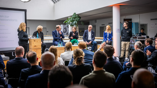 Im Vordergrund des Bildes ist ein Teil eines Publikums von hinten zu sehen. Vorne auf einer Erhöhung befinden sich FH-Rektorin Prof. Tamara Appel, IHK-Hauptgeschäftsführer Stefan Schreiber, die Geschäftsführerin der Wirtschaftsförderung Heike Marzen, TU-Rektor Prof. Manfred Bayer und ISM-Vizepräsidentin Prof. Audrey Mehn.