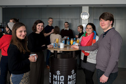 The picture shows several people standing around a bar table and smiling into the camera. There are other people standing around bar tables in the background.