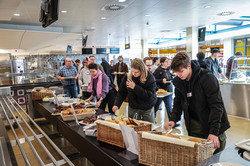 Menschen stehen an einem Buffet in einer Mensa und füllen ihre Teller mit Essen.