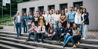 Gruppenfoto auf einer grauen Treppe. Im Hintergrund ein kleines, graues Gebäude der RUB.