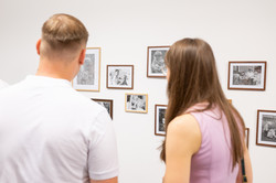 Das Bild zeigt zwei Personen von hinten, die vor einer weißen Wand mit vielen kleinen eingerahmten Kunstwerken von Studierenden stehen.
