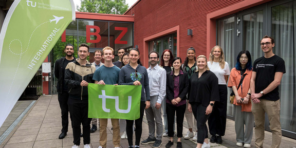Eine Gruppe von Menschen steht vor dem Internationalen Begegnungszentrum (IBZ). Drei Menschen im Vordergrund halten eine Fahne mit dem Logo der TU Dortmund.
