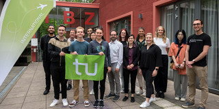Eine Gruppe von Menschen steht vor dem Internationalen Begegnungszentrum (IBZ). Drei Menschen im Vordergrund halten eine Fahne mit dem Logo der TU Dortmund. 