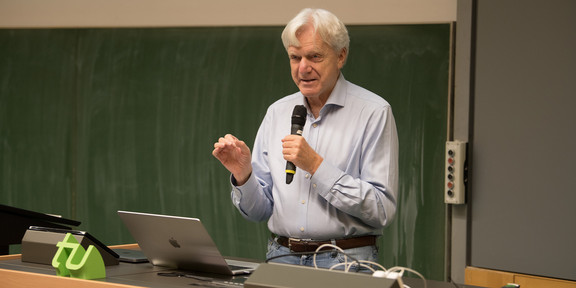Andreas von Bechtholsheim, ein Mann mit grauen Haaren und hellblauem Hemd, steht mit einem Mikrofon in der Hand vor einer Tafel und hält einen Vortrag.