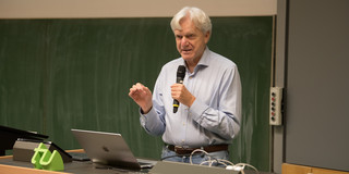 Andreas von Bechtholsheim, ein Mann mit grauen Haaren und hellblauem Hemd, steht mit einem Mikrofon in der Hand vor einer Tafel und hält einen Vortrag.