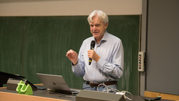 September Andreas von Bechtholsheim, ein Mann mit grauen Haaren und hellblauem Hemd, steht mit einem Mikrofon in der Hand vor einer Tafel und hält einen Vortrag.