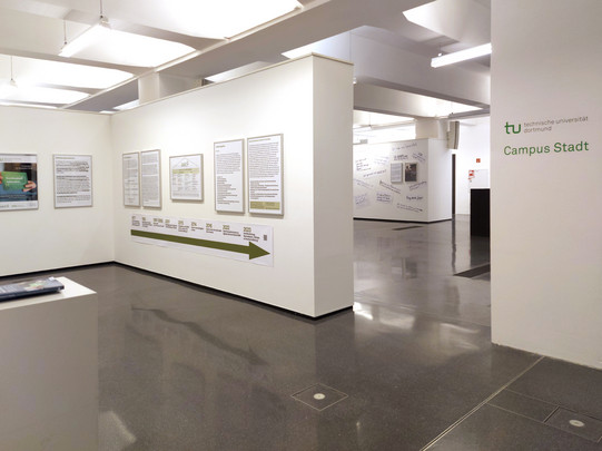Foto: Heller Ausstellungsraum mit weißen Wänden, mehreren gerahmten Infotafeln und einer Zeitstrahl-Grafik, reflektierendem Boden und der Aufschrift "Campus Stadt" der Technischen Universität Dortmund am Eingang.